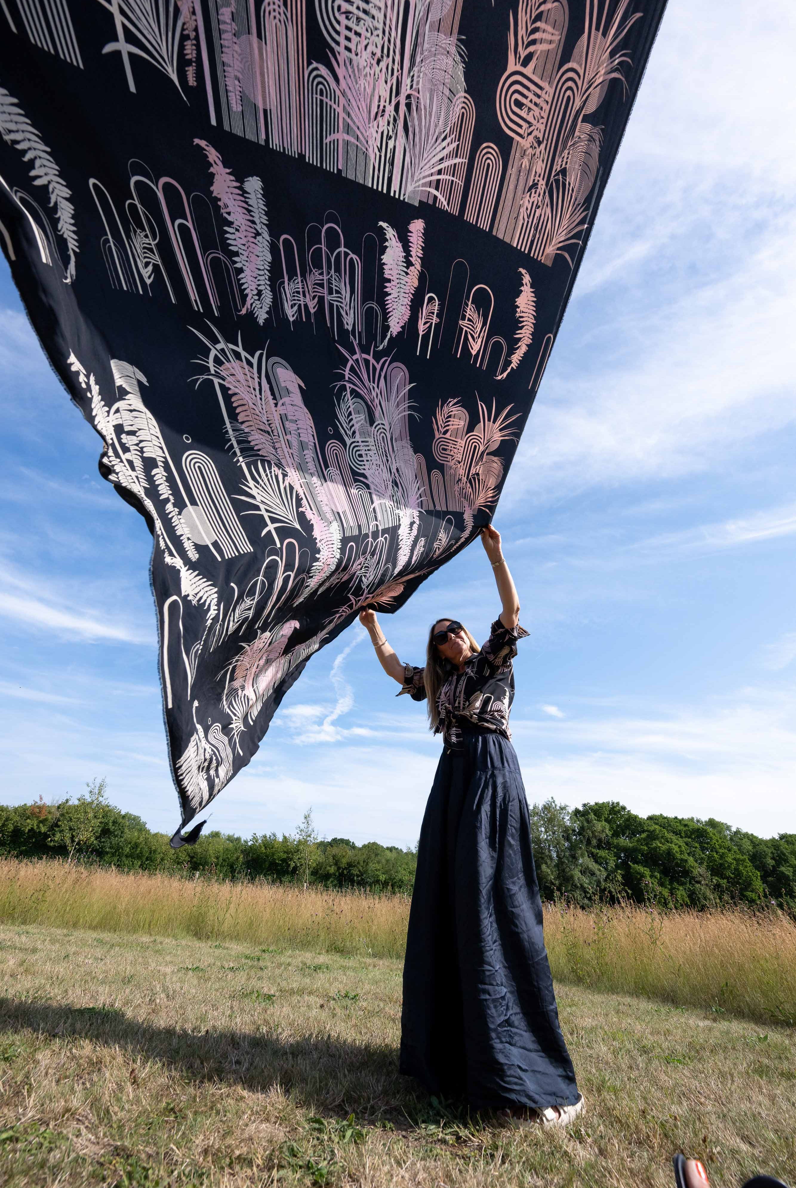 Person holding up a large black fabric with white and pink designs against a blue sky.