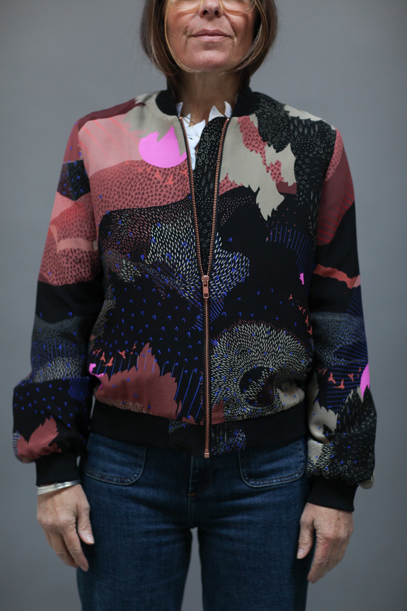 A woman wearing a silk bomber jacket in black and pink with a landscape design and bright pink suns