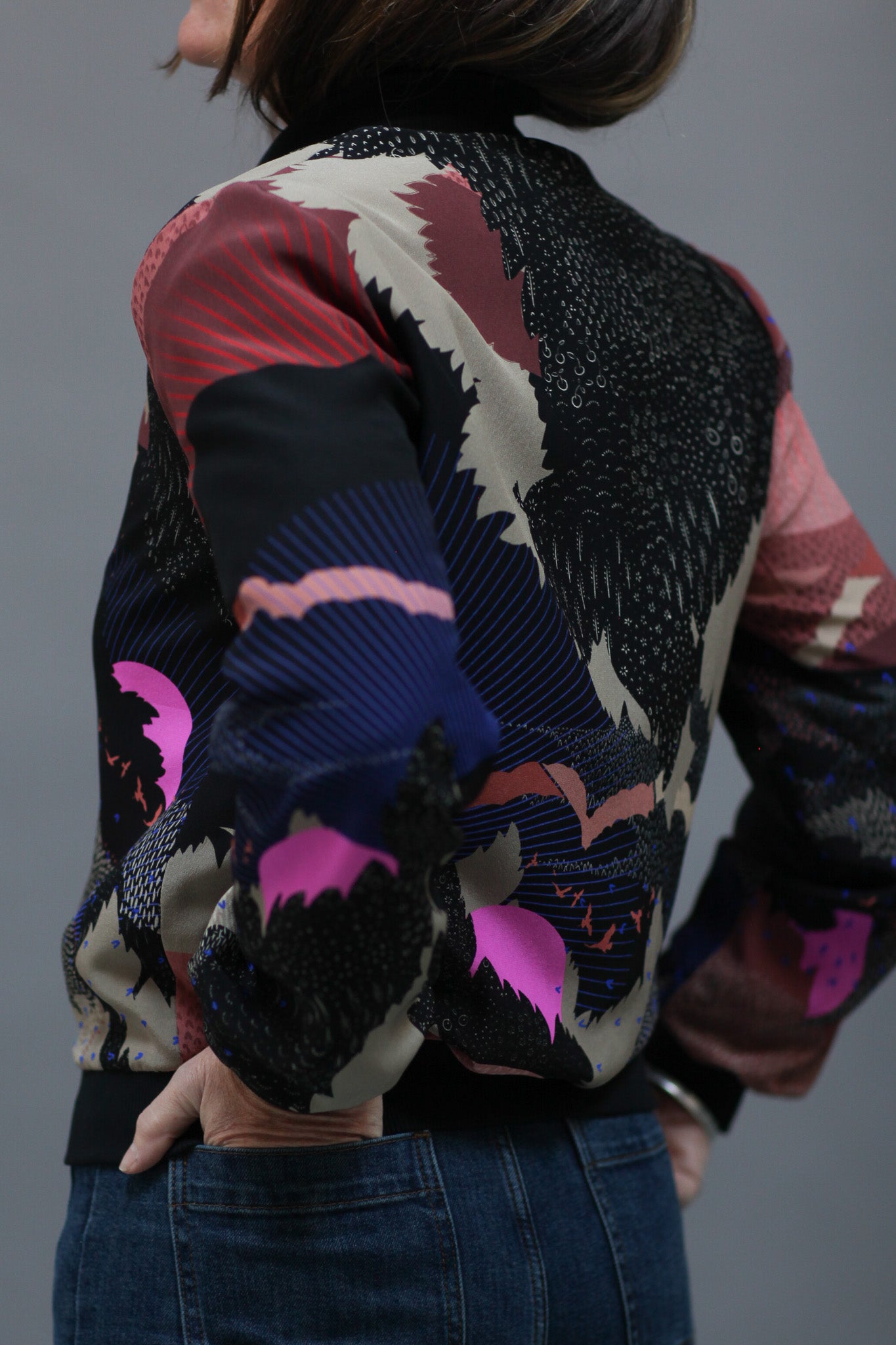 A woman wearing a silk bomber jacket in black and pink with a landscape design and bright pink suns
