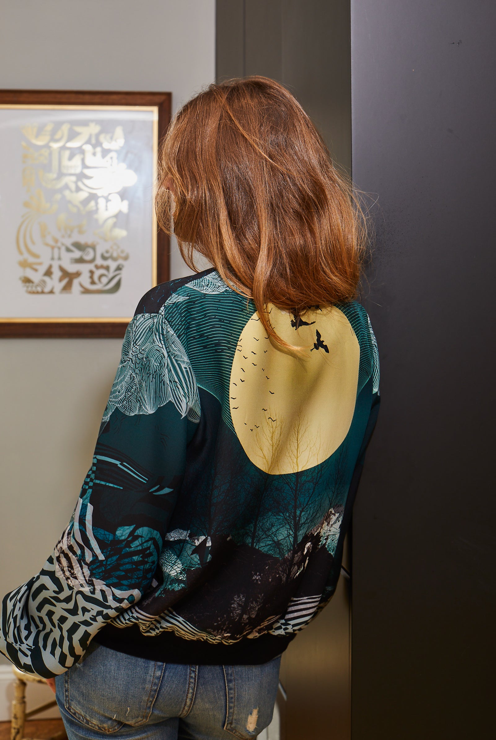 A woman's back wearing a silk bomber jacket with a large yellow moon and birds with blue landscapes