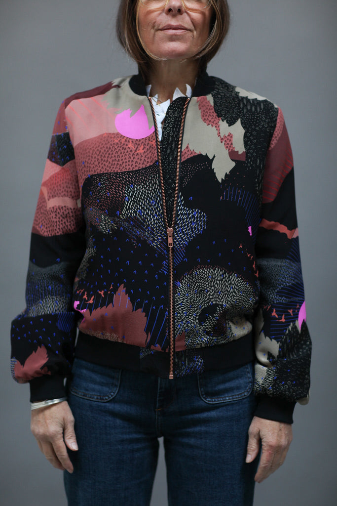 A woman wearing a silk bomber jacket in black and pink with a landscape design and bright pink suns 