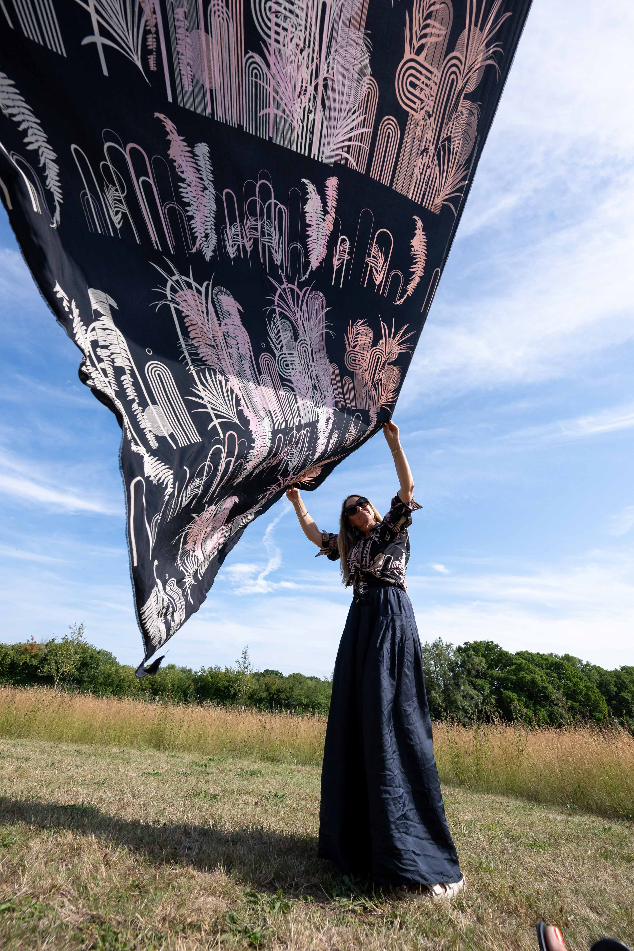 Person holding up a large black fabric with white and pink designs against a blue sky.