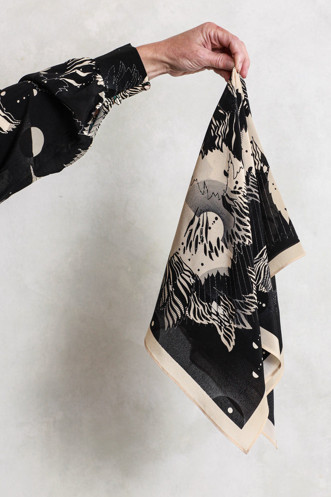 A silk scarf being held against a white wall. It is an abstract floral design in cream and black.