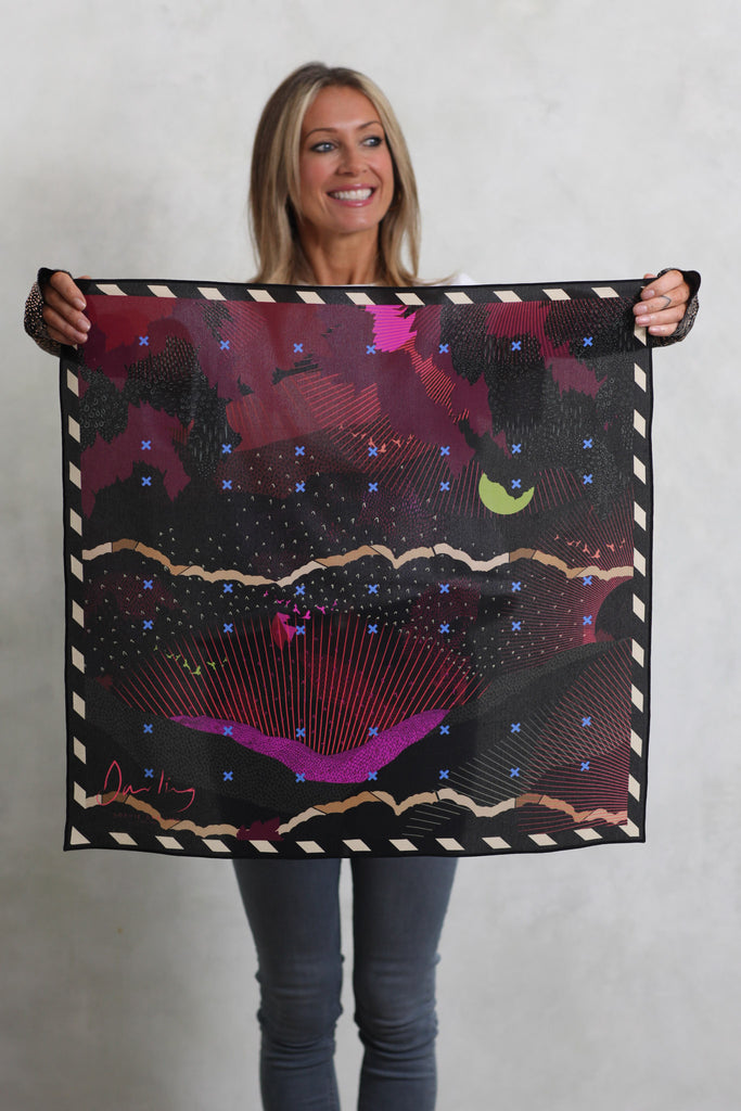 A woman holding a silk scarf with a landscape design in pinks, blacks and a lime green moon