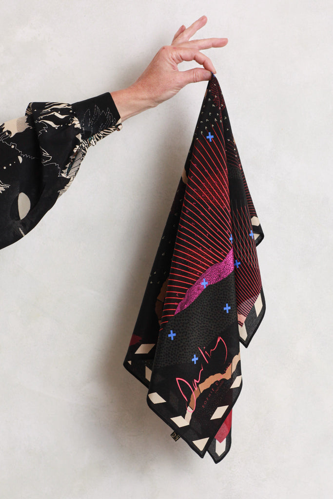 A silk scarf with a landscape design in pinks, blacks and greens being held against a white wall