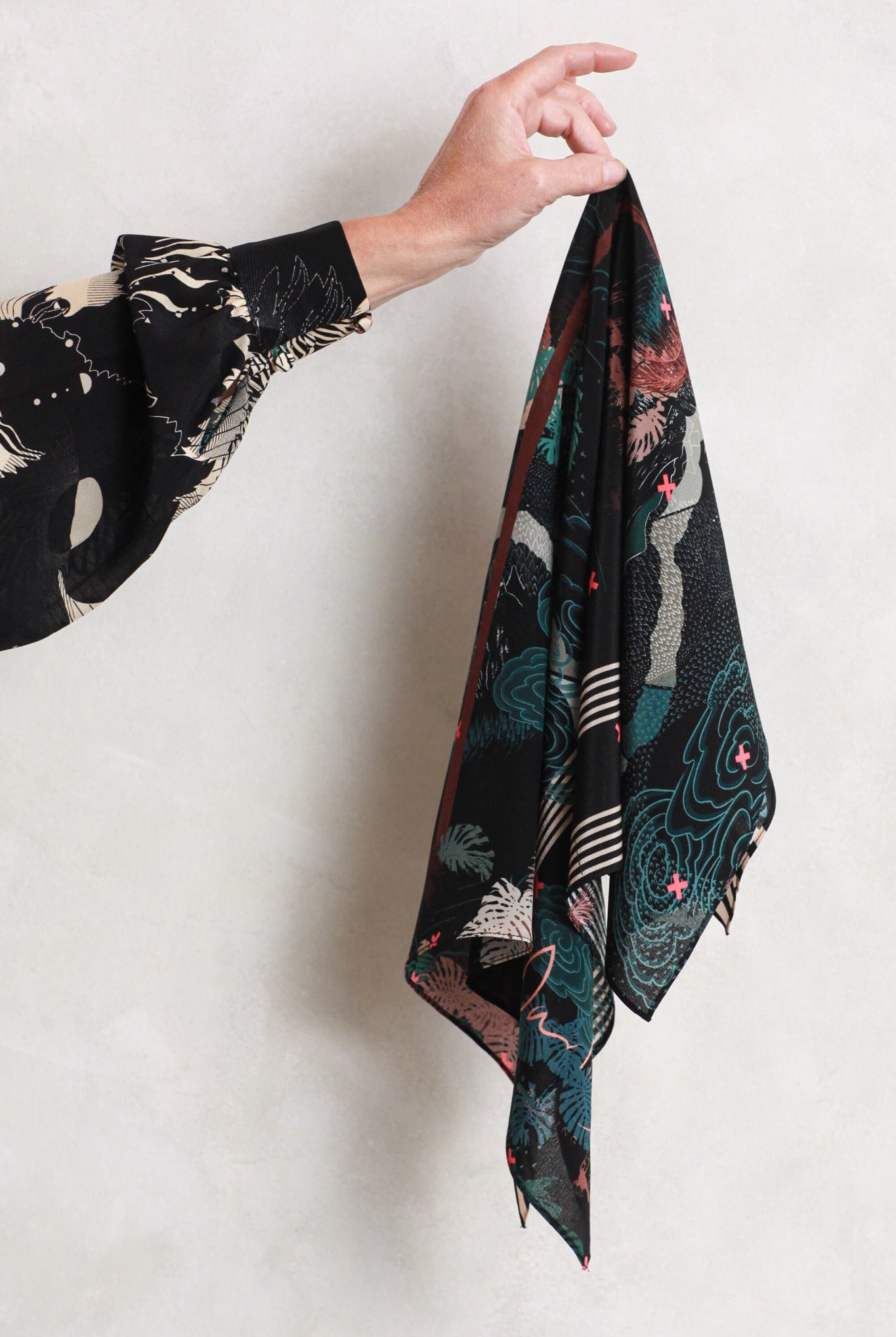 A silk scarf being held against a white wall. It has a tropical design in blacks, greens and pinks