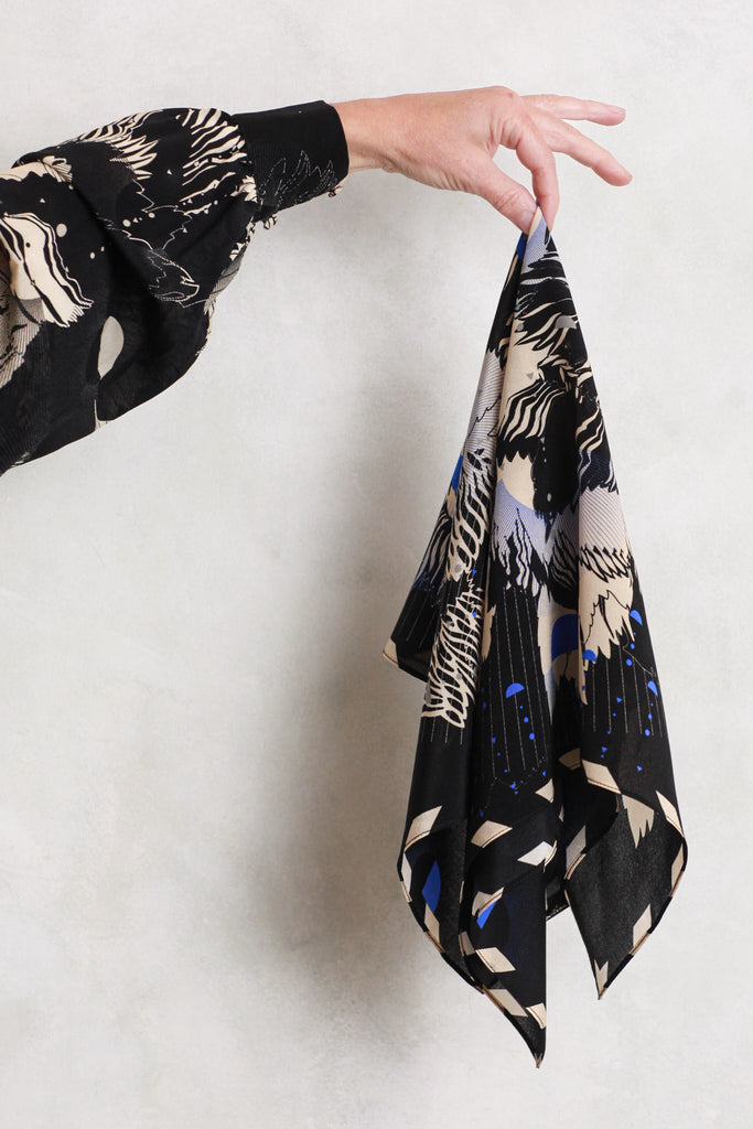 A silk scarf being held against a white wall. It is an abstract floral design in cream and navy blue