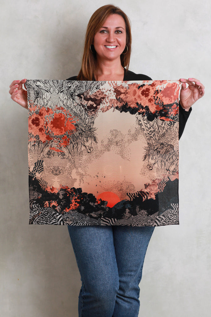 A woman holding a silk scarf with a sunset design in oranges and blacks