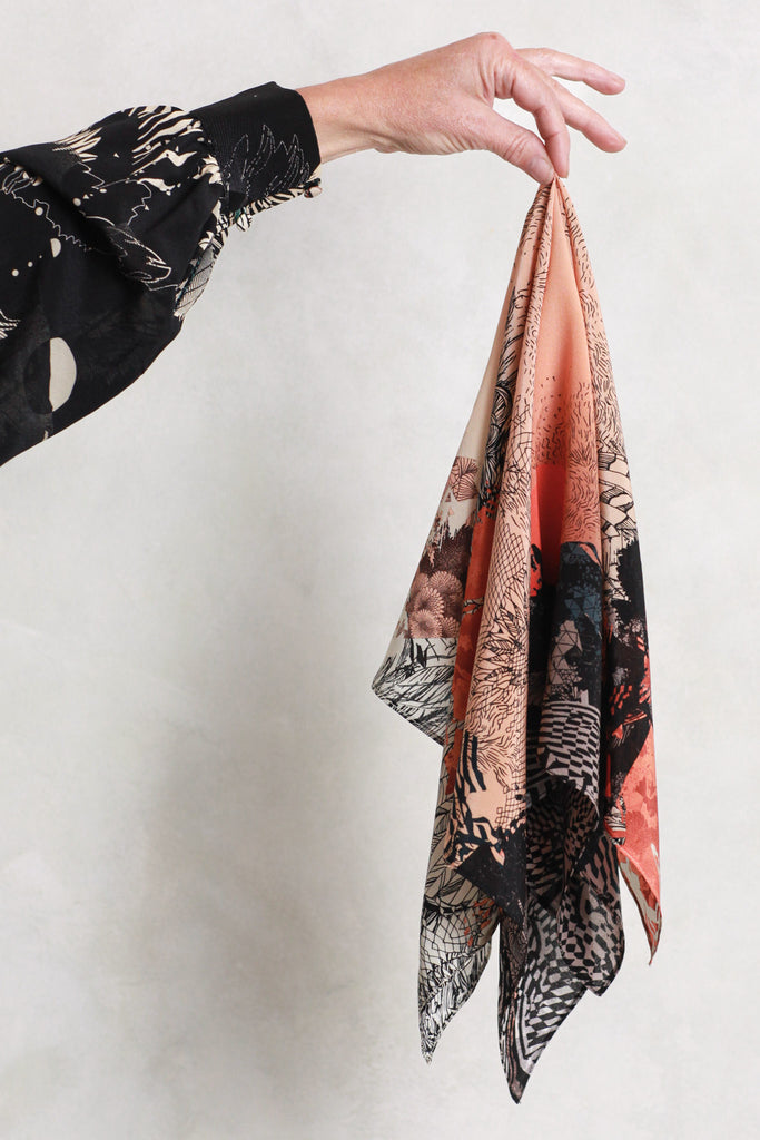 A silk scarf being held against a white wall. It has a sunset design in oranges and blacks