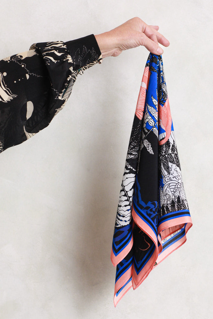 A silk scarf being held against a white wall. It has a wave and birds design in blue and coral