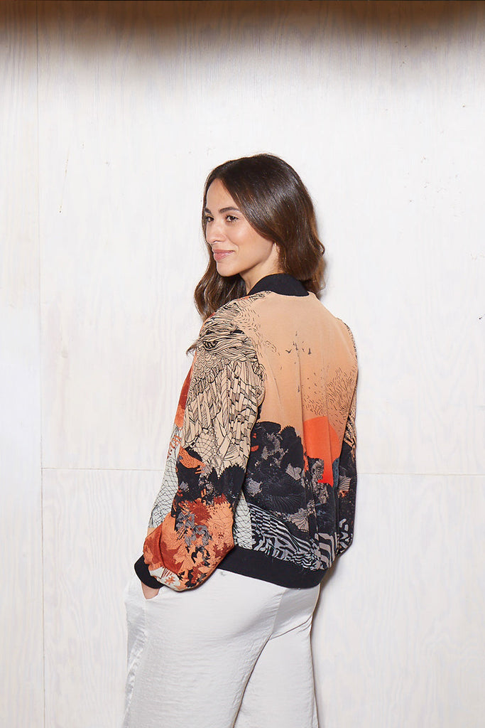 Woman wearing a Silk bomber jacket in orange and black with sunrise design in front of white wall