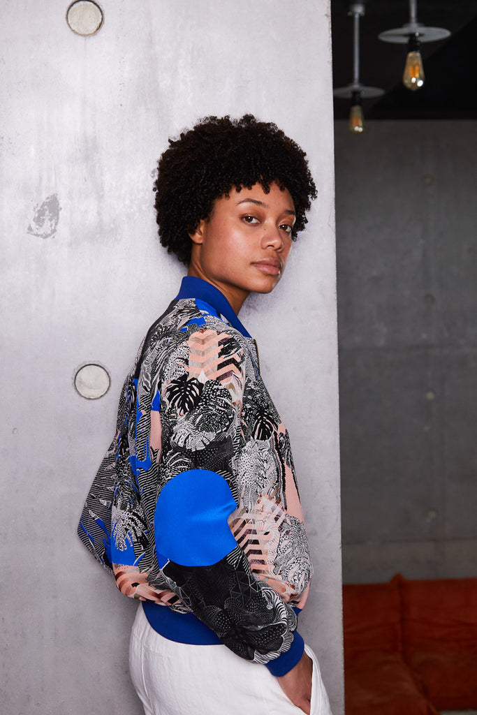 A woman side on wearing a silk bomber jacket in blue, pink and monochrome tropical print