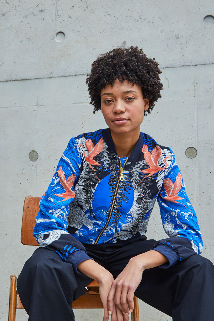 A female model sat down wearing a blue printed silk bomber jacket with coral birds on